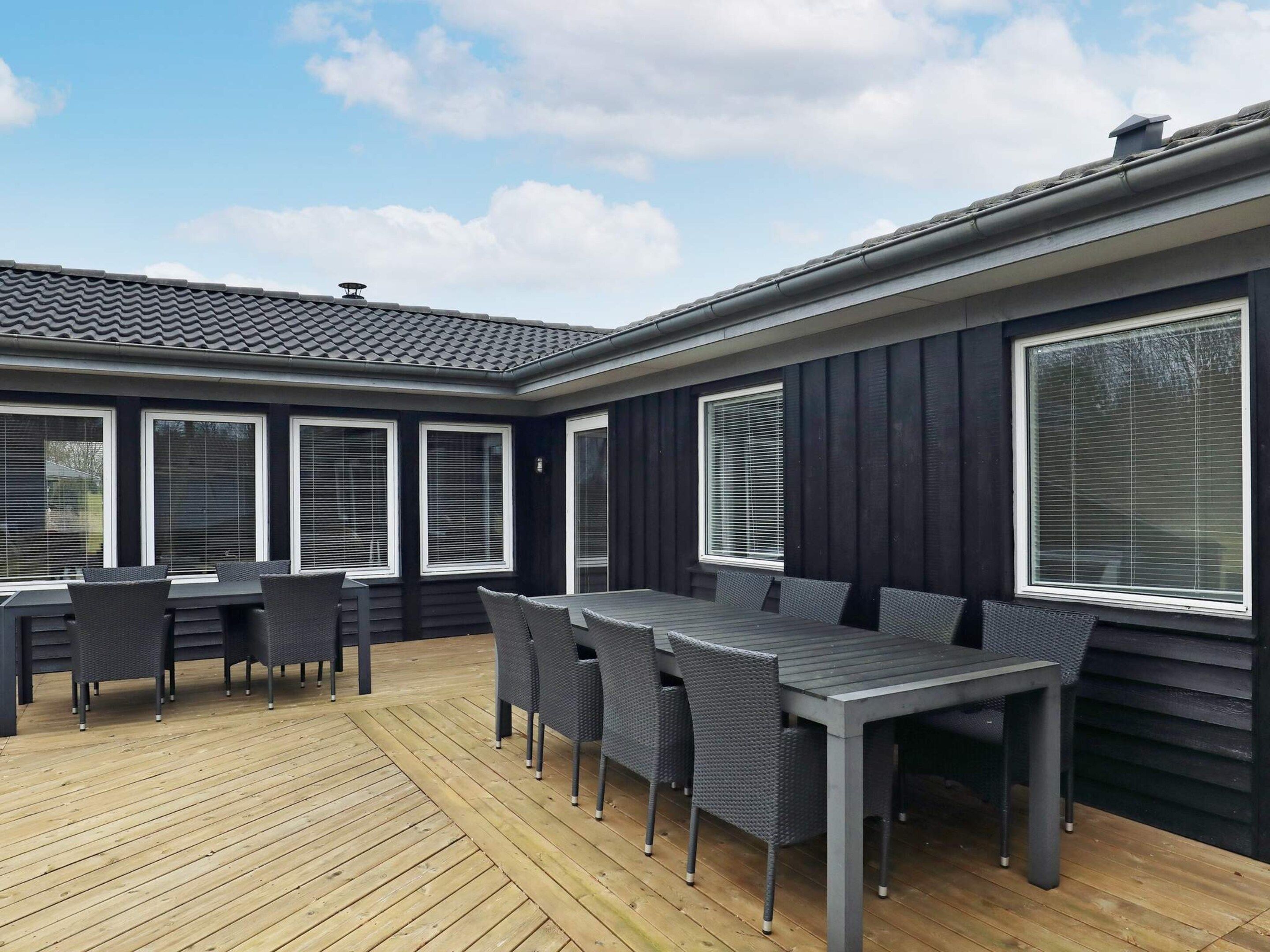 A covered outdoor patio with a wooden deck, a dining table with chairs, and large windows with blinds.