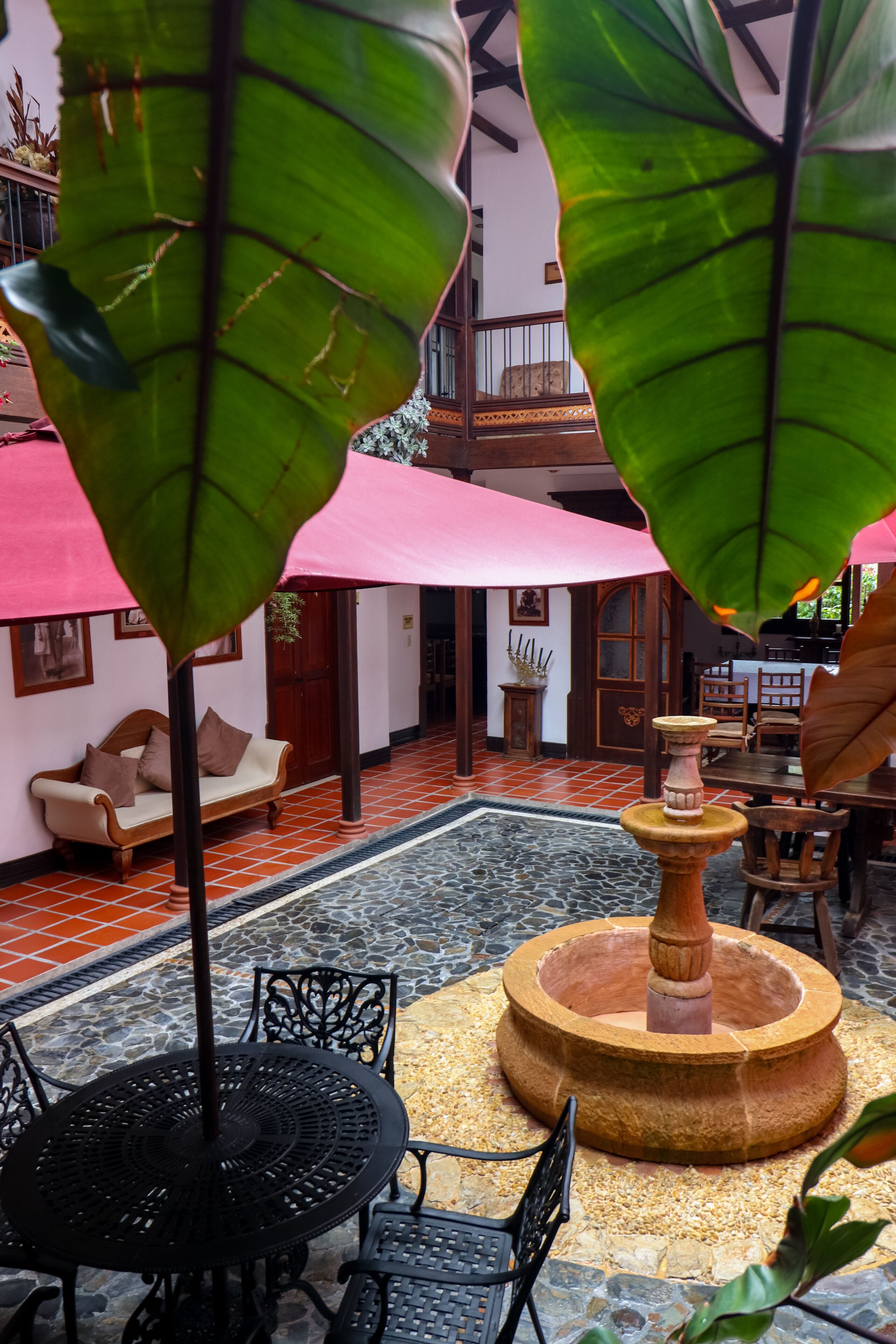 Un cortile con pavimento in pietra, un tavolo rotondo e sedie, circondato da grandi foglie verdi e un ombrello rosa.