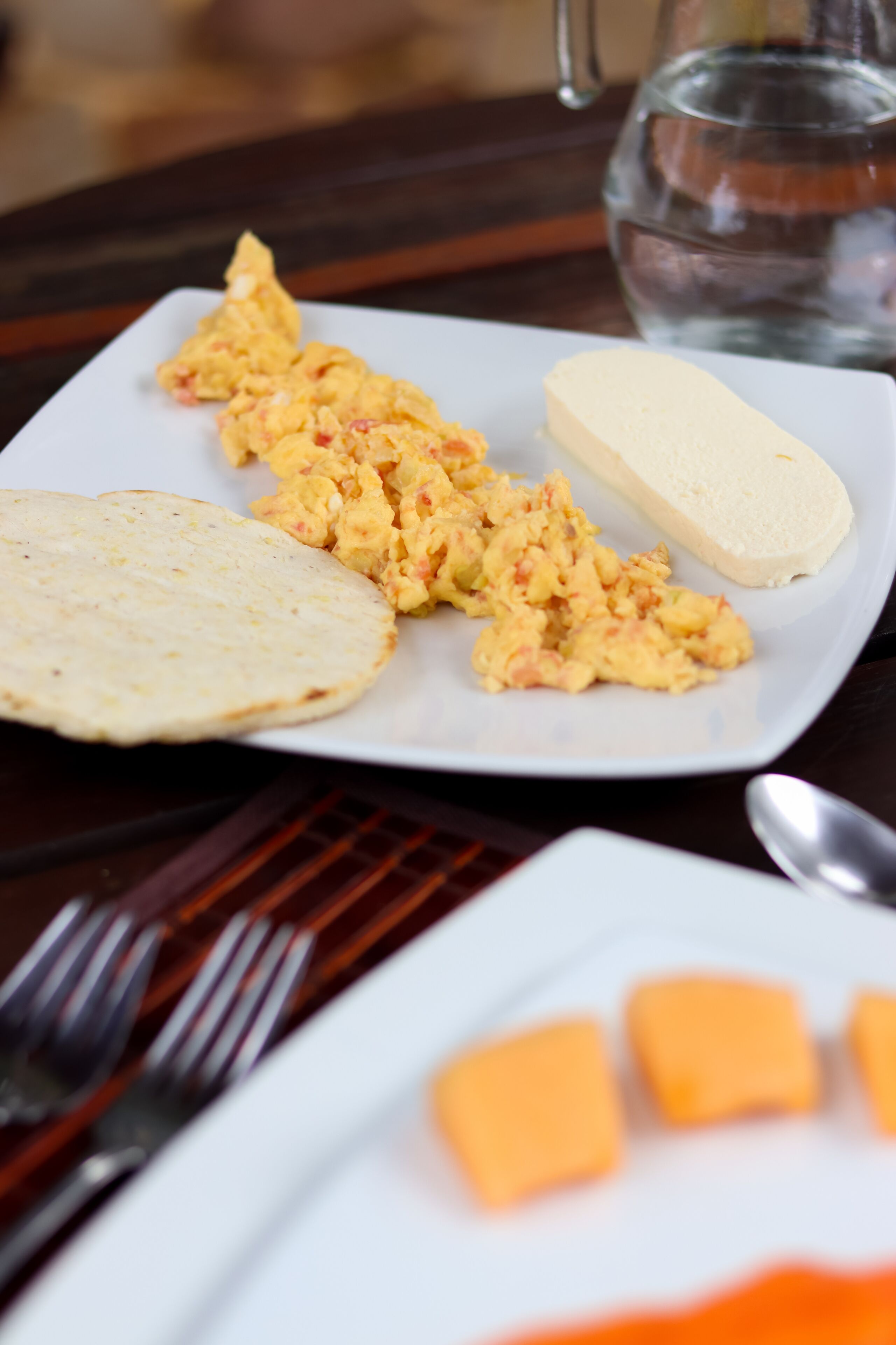 A white plate with scrambled eggs, a slice of bread, and a glass of water on a wooden table.