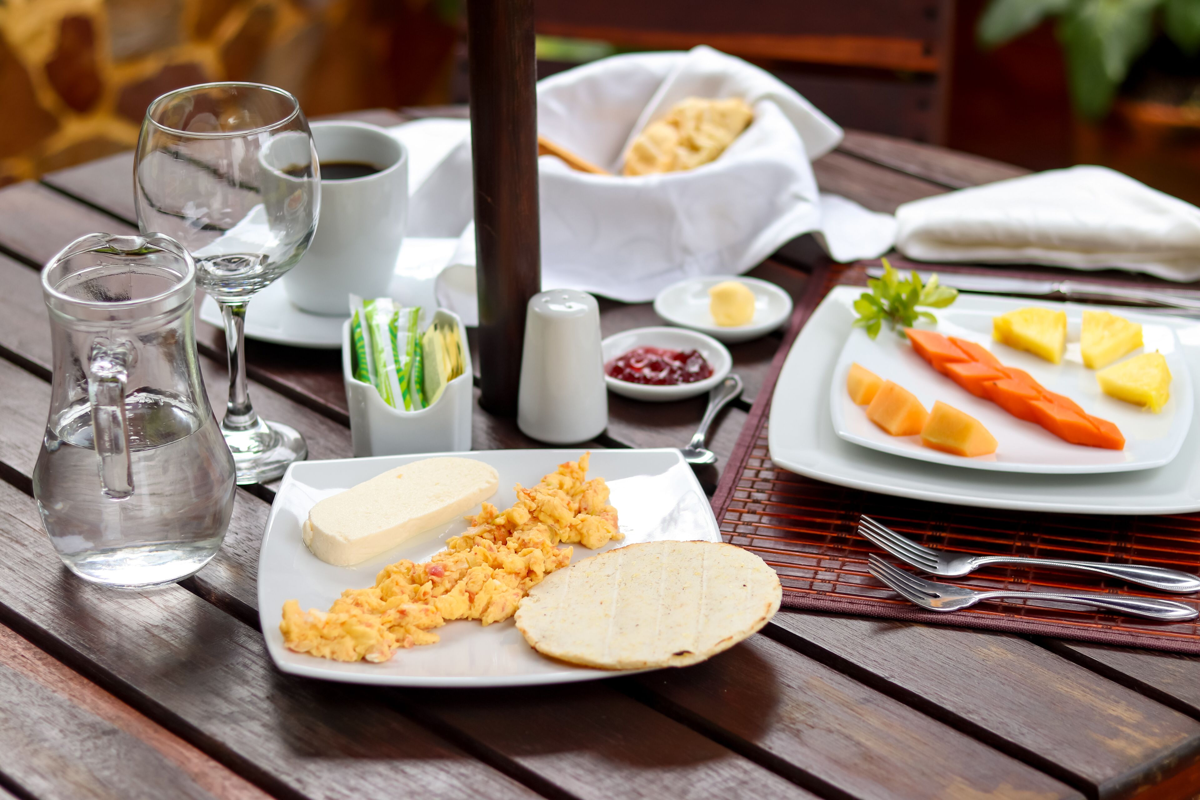 Una mesa de madera con alimentos para el desayuno, incluyendo huevos revueltos, tostadas y una fuente de fruta.