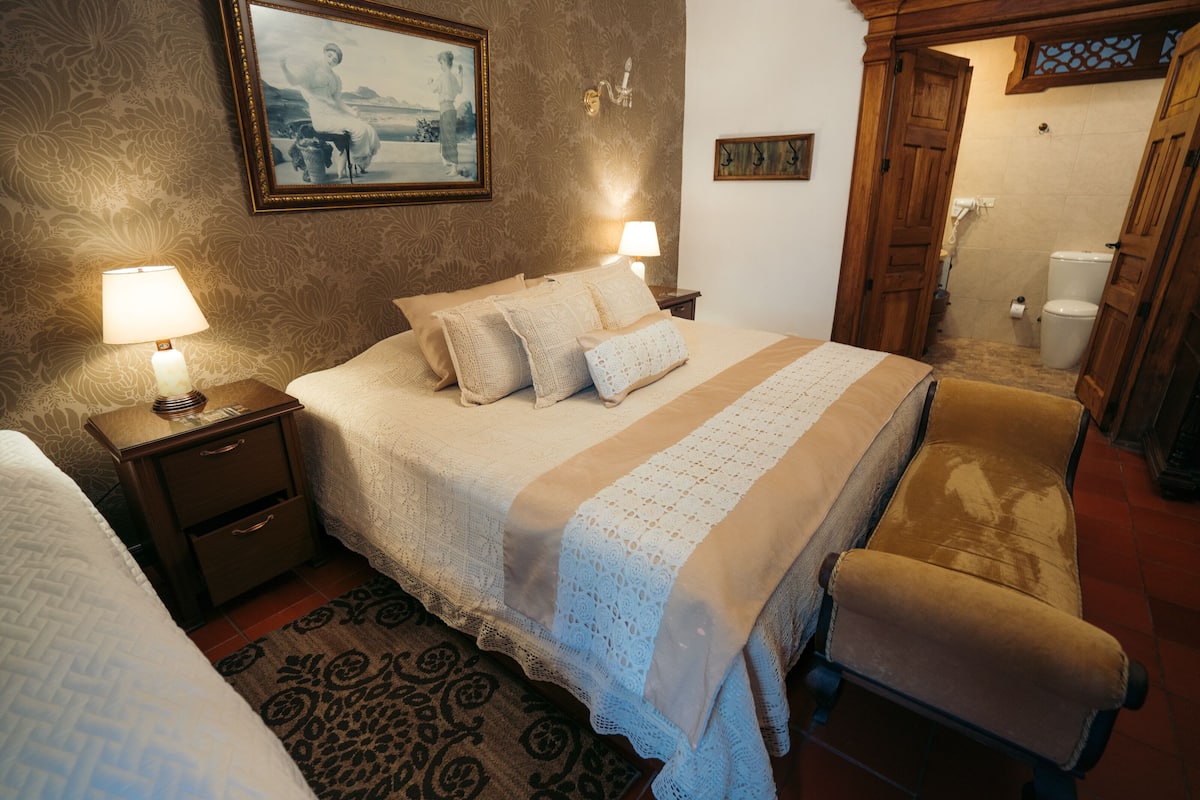 Een keurig opgemaakt slaapkamer met een bed, twee nachtkastjes met lampen, een stoel, en een zichtbare badkamer door een open deur.