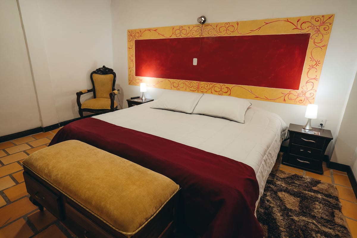 Een hotelkamer met een groot bed, een nachtkastje met een lamp, een stoel en een decoratief hoofdeinde.