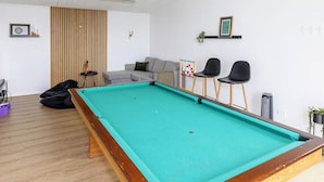 Une table de billard dans une pièce où se trouvent un canapé, deux fauteuils, un jeu de fléchettes et une petite plante.