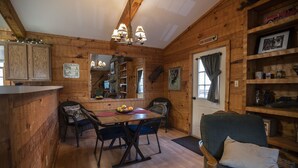 Interior de una cabaña de madera con una mesa de comedor, sillas, una araña, chimenea y una estantería.