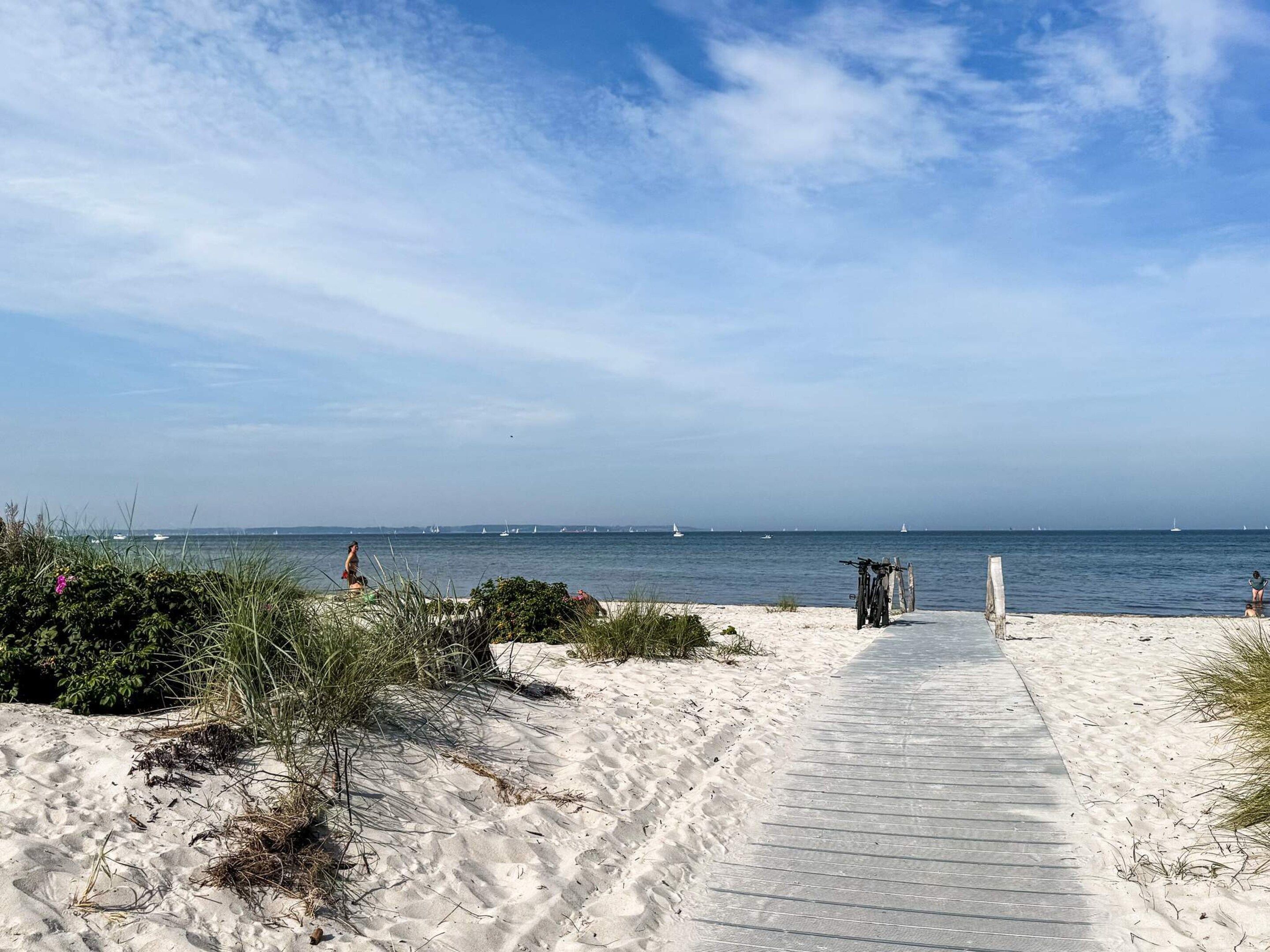 Una playa arenosa con un paseo marítimo de madera que lleva al agua, barcos a lo lejos y vegetación escasa.