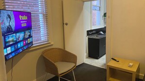 A room with a television displaying a streaming service interface, a wicker chair, a small table, and a door leading to a kitchen area.