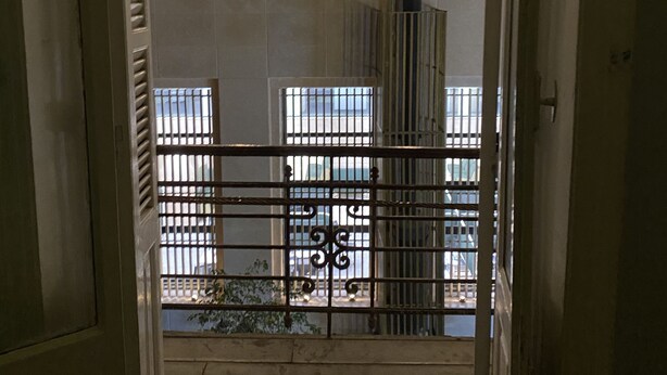 Blick durch ein Metallgitter in ein Gebäude mit Fliesenboden, einer Treppe und einem Fenster mit Jalousien.