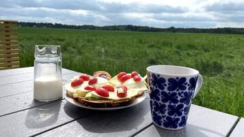 Una mesa con un plato de comida, un vaso de leche y una taza azul con motivos florales, sobre un fondo de campo verde y cielo nublado.