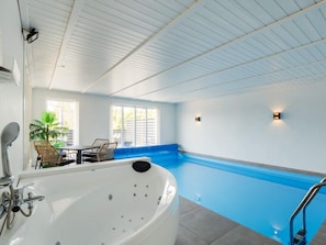 En moderne indendørs swimmingpool med blå kant, et spabad og et loungeområde med stole og et bord.