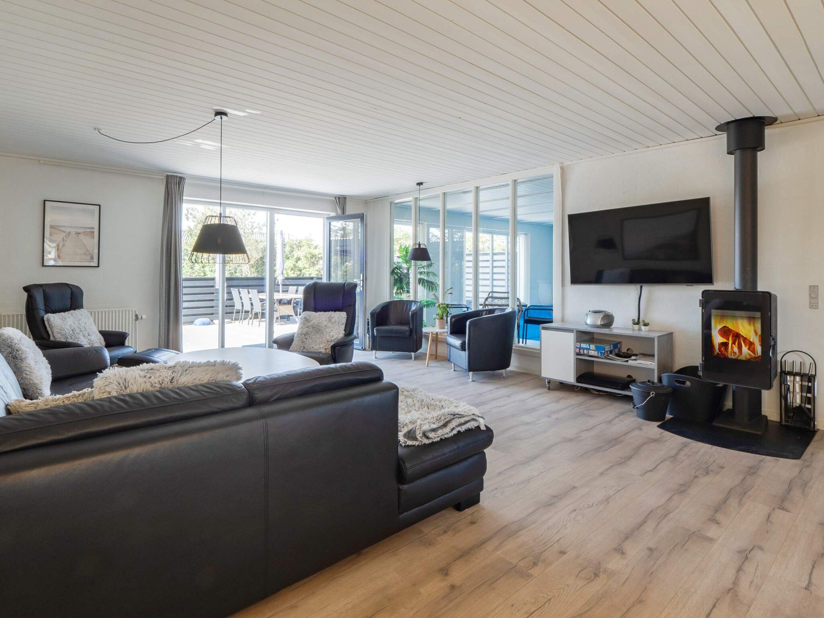 A modern living room with a black leather sofa, a flat-screen TV, a wood-burning stove, and large windows.