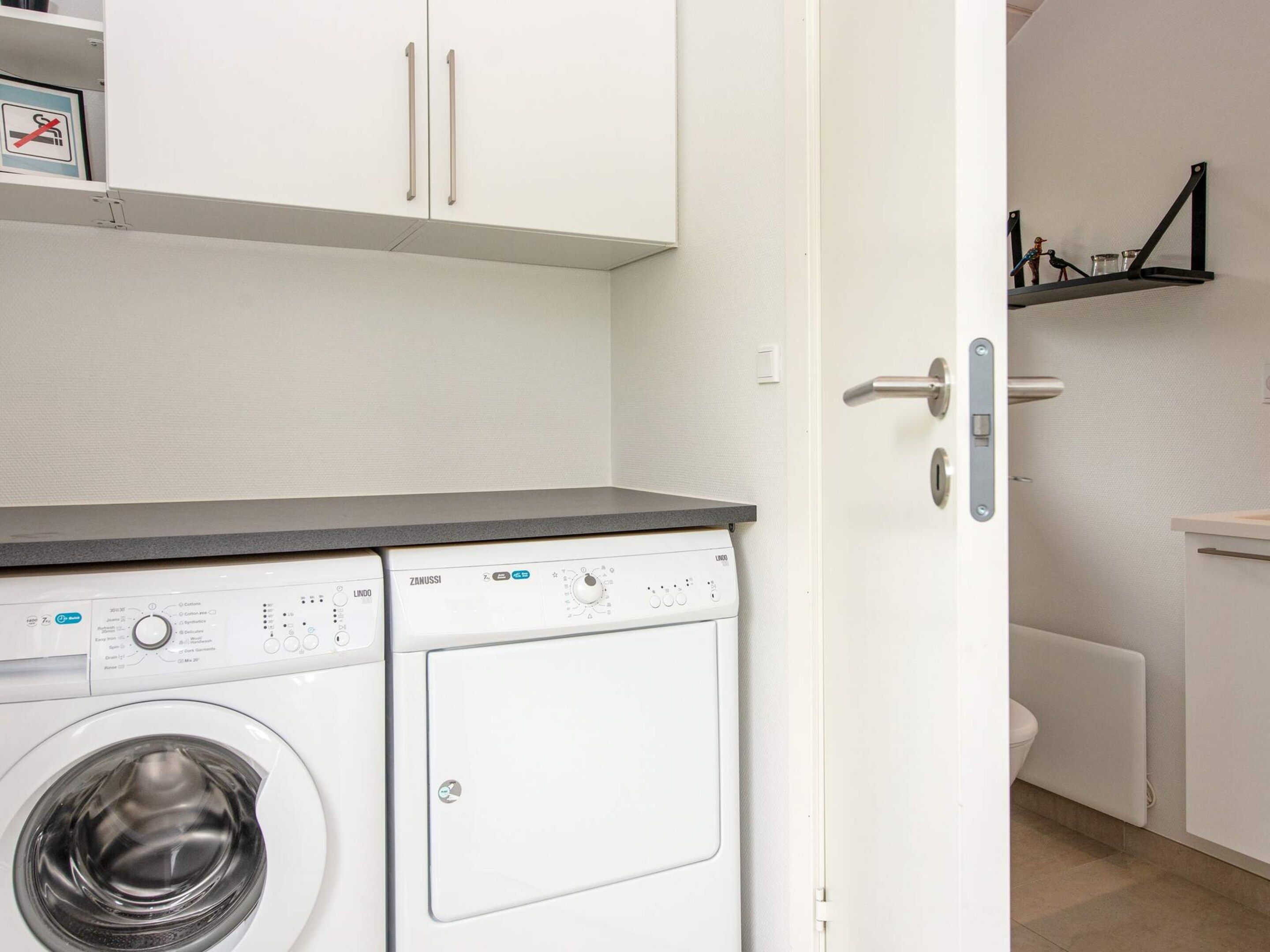 A laundry area with a front-loading washing machine, a white cabinet with a metal handle, and a shelf with a no-smoking sign.