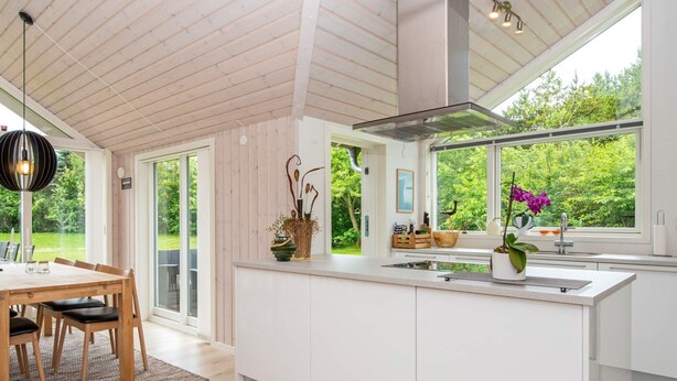 Een moderne keuken met een groot eiland, een houten eettafel en stoelen, grote ramen met uitzicht op groen.