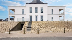 A modern two-story building with a staircase leading to the entrance, situated on a sandy terrain with a clear sky. - 4 Star Holiday Home in Ringkobing (Ringkobing)