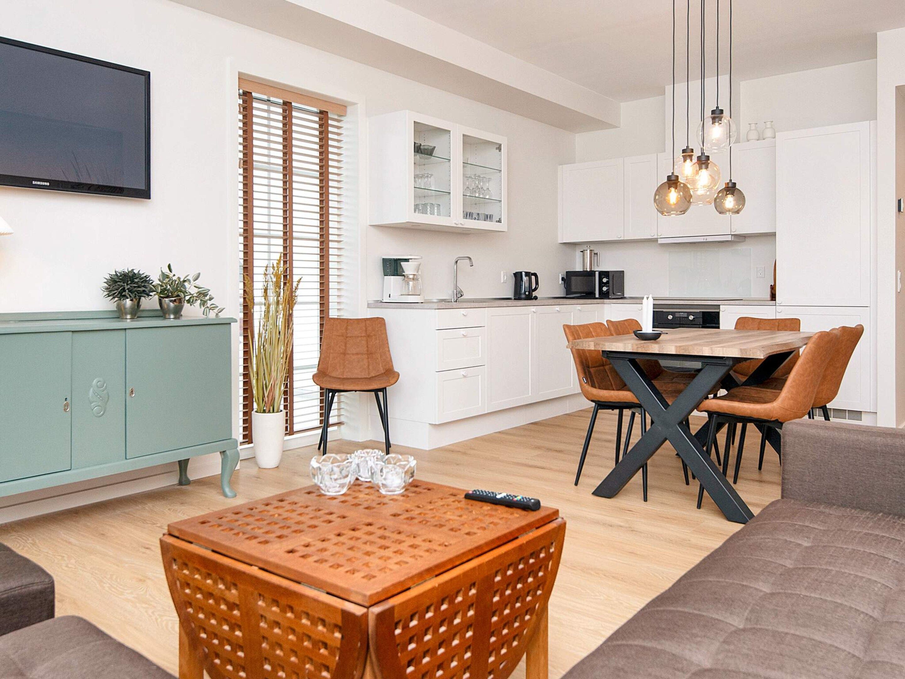 A modern kitchen with white cabinets, a dining table with chairs, a TV mounted on the wall, and a potted plant.
