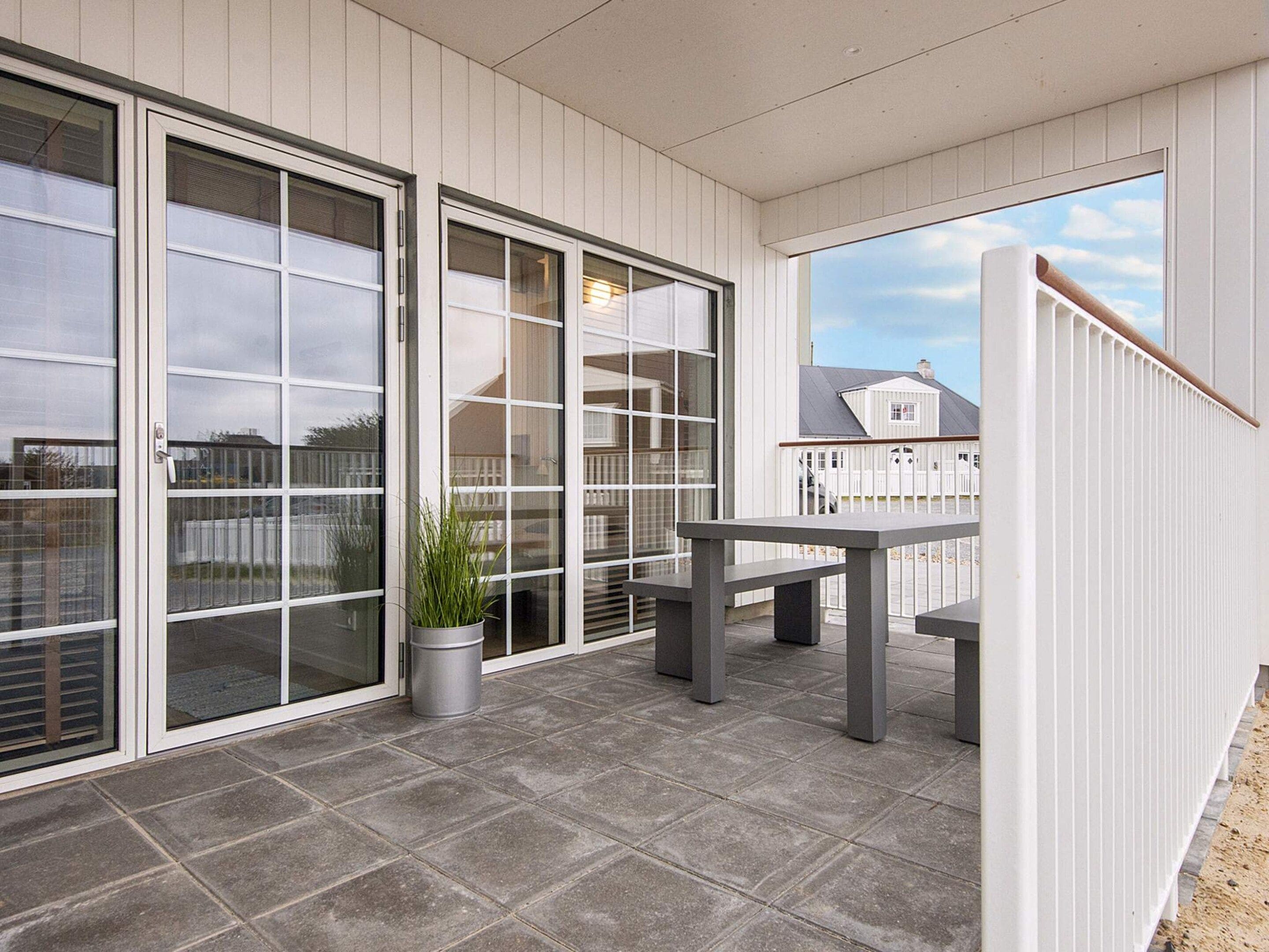 A covered patio area with a table and chairs, a potted plant, and a view of a white fence and houses in the background.
