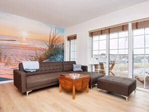 A modern living room with a large window, a brown sofa, a wooden coffee table, and a view of the beach.