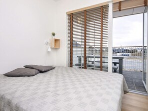 Un dormitorio con cama, mesita de noche, lámpara y vista a un estacionamiento a través de una ventana con persianas.