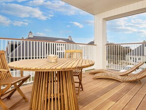 Una terraza de madera con una mesa redonda y sillas, baranda blanca y vistas a casas al fondo.
