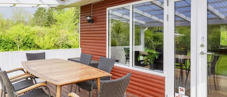 A covered patio with a wooden table and chairs, a glass door leading to a garden, and a red exterior wall.