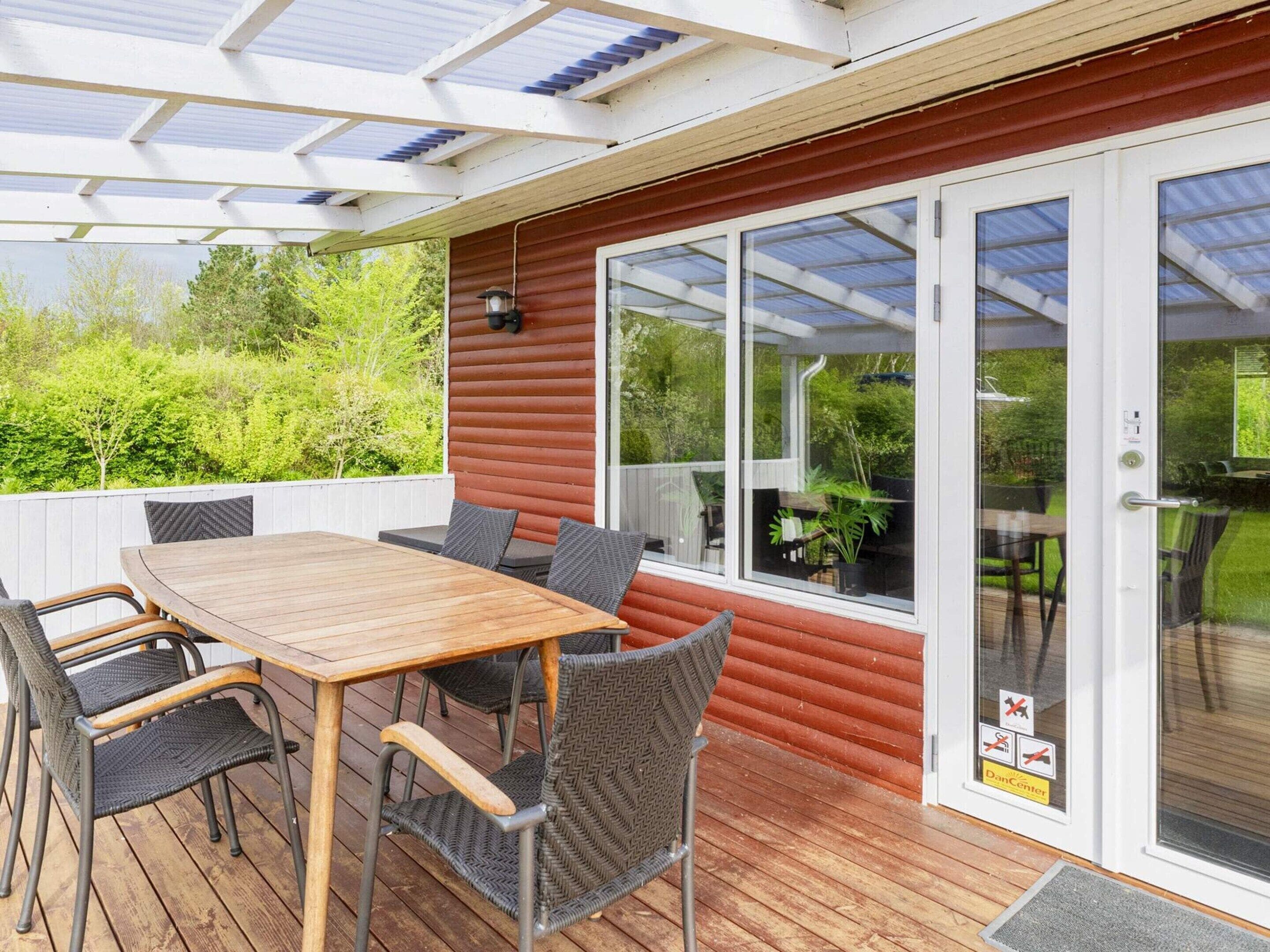 Un patio al aire libre con mesa y sillas de madera, una puerta de cristal que da a un jardín y una estructura de techo con marco de metal.