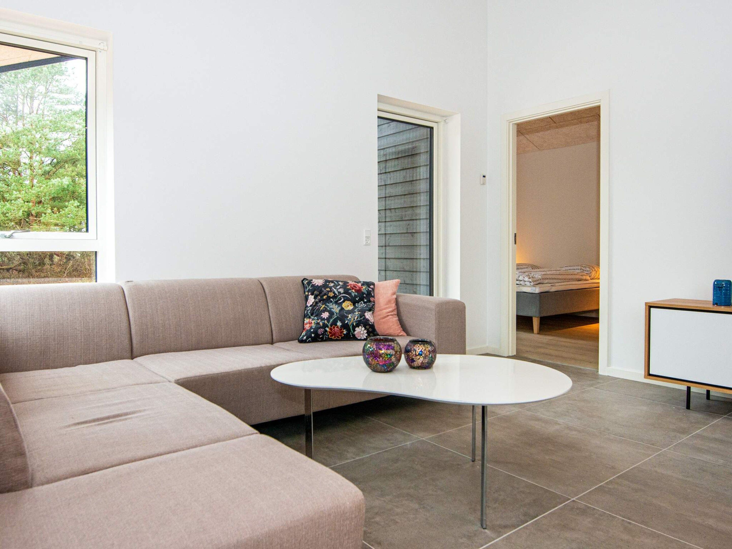 A modern living room with a beige sectional sofa, a small white coffee table, and a view of a bedroom through an open door.
