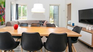 A modern dining area with a wooden table, black chairs, a sofa with cushions, a white pendant light, and a television on a stand.