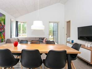 A modern dining area with a wooden table, black chairs, a sofa with cushions, a white pendant light, and a television on a stand.