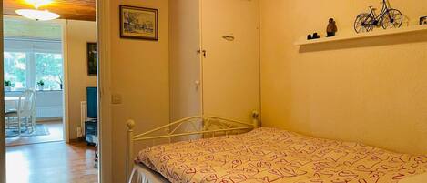 A bedroom with a bed, a wardrobe, a door, a shelf with items, and a window with blinds.