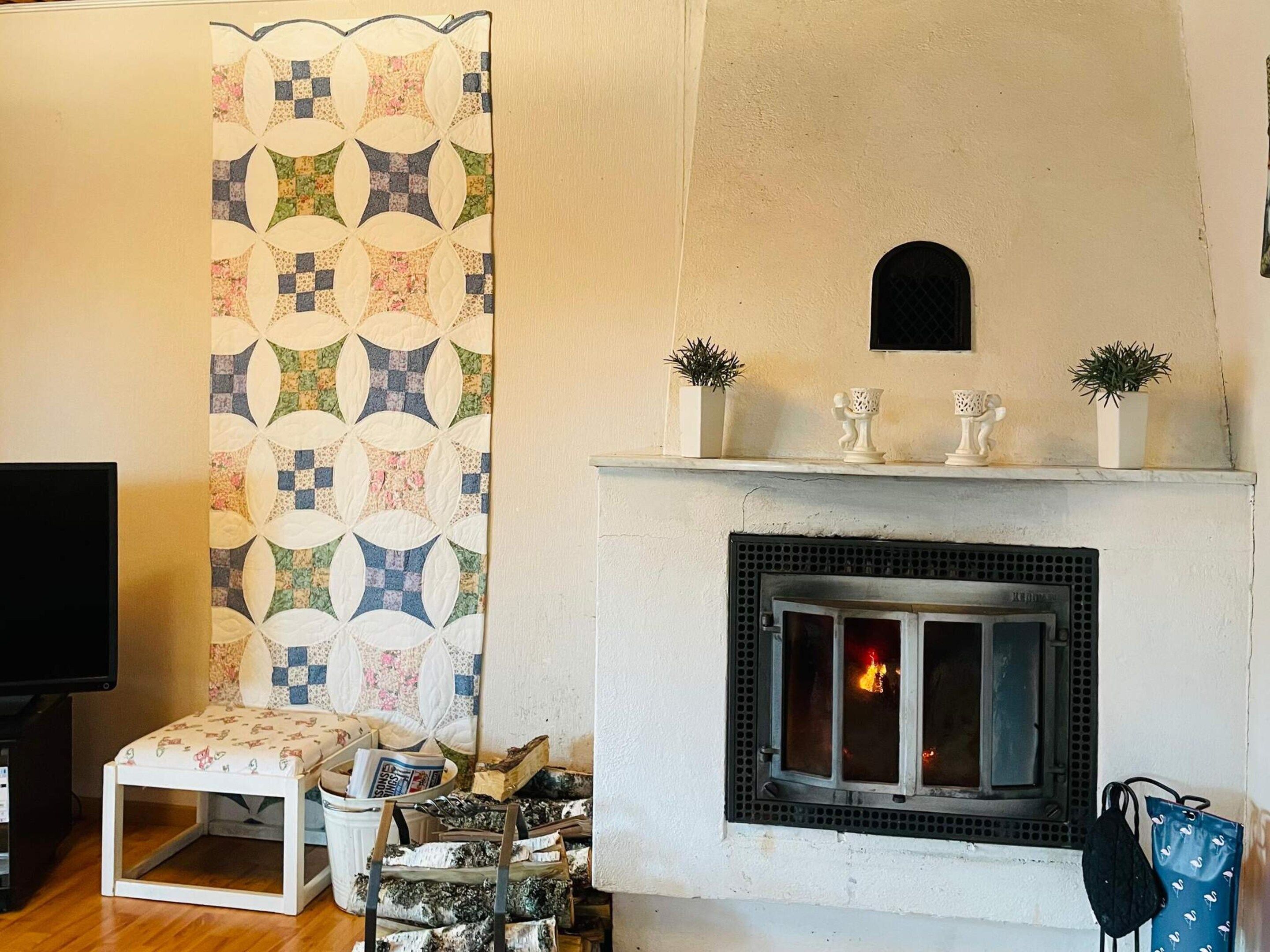 A living room with a fireplace, a television, a small table with newspapers, and a patterned wall hanging.