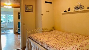 Une chambre avec un lit simple, une armoire, une petite étagère avec des objets décoratifs et une porte donnant sur un balcon.