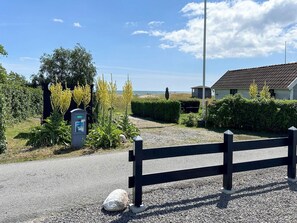 Une allée de gravier mène à une maison entourée d’une clôture noire, avec un coffret électrique à proximité, et offrant une vue sur la mer.
