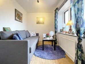 Un salon avec un canapé gris, une petite table sur laquelle se trouve un ventilateur, un tapis bleu et une fenêtre avec des rideaux.