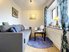 A living room with a grey sofa, a small table with a fan, a blue rug, and a window with curtains.
