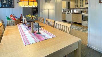 A dining area with a wooden table, chairs, a striped table runner, a hanging lamp, and a view into the kitchen.