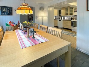 A dining area with a wooden table, chairs, a striped table runner, a hanging lamp, and a view into the kitchen.