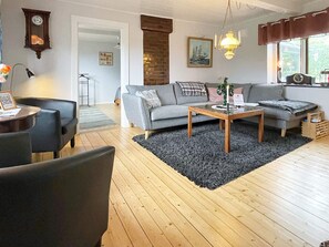 Ein Wohnzimmer mit einer Couch, einem Couchtisch und Sesseln. Es gibt einen Kamin, eine Uhr und ein Fenster mit Vorhängen.