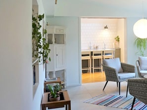Uma sala de estar moderna com cozinha ao fundo, com móveis de vime, um tapete listrado e uma planta em vaso.