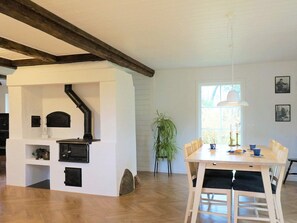 Une pièce dotée d’un poêle à bois encastré, d’une table à manger avec des chaises, d’une plante en pot et d’une fenêtre laissant entrer la lumière naturelle.