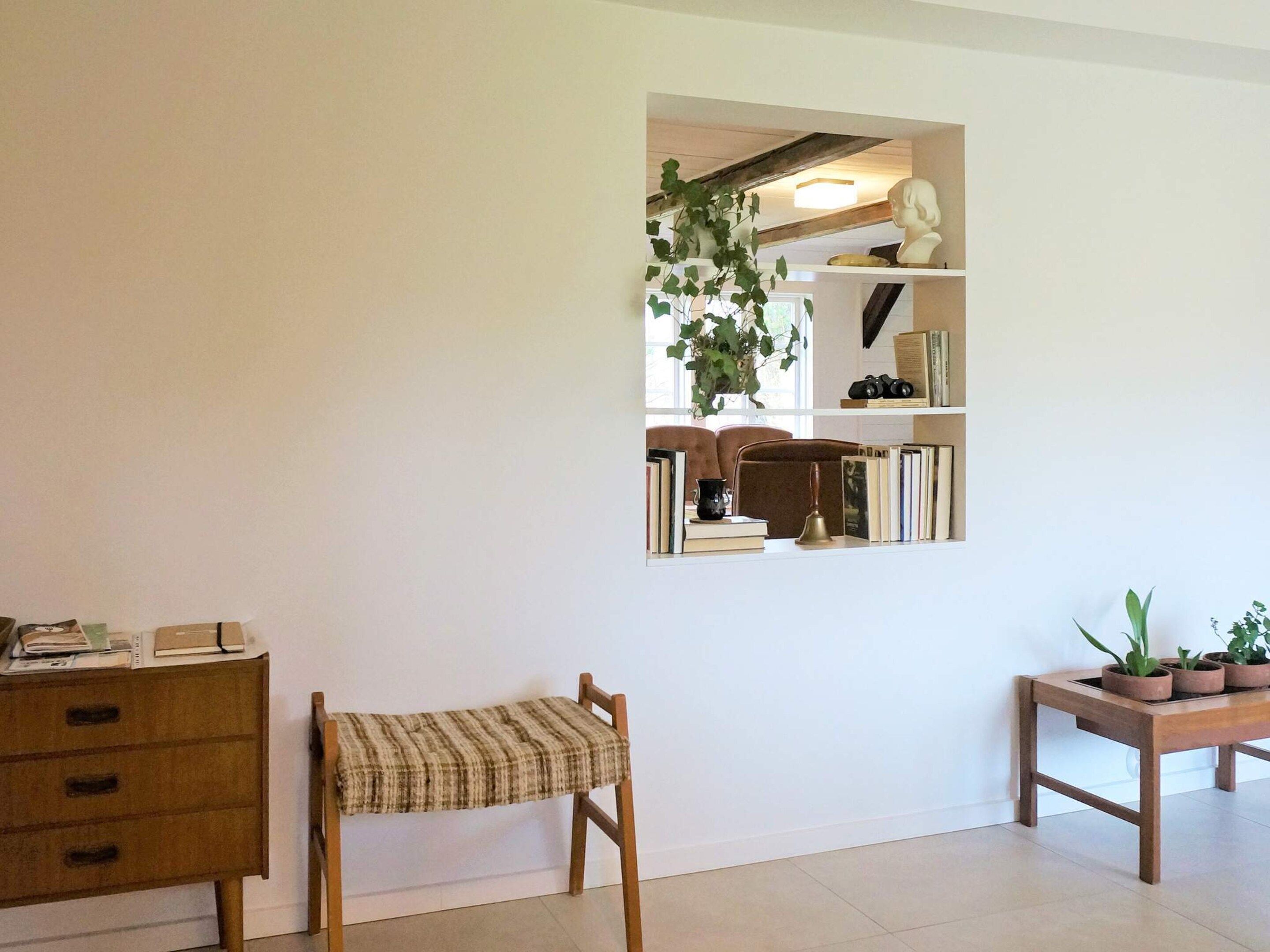 A room with a wooden chair, a small table with potted plants, a bookshelf, and a mirror reflecting the room.