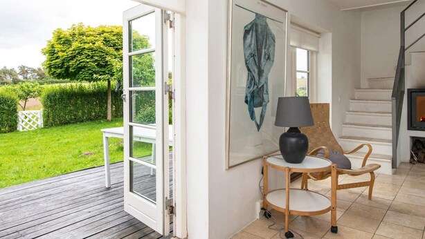 A room with a wooden deck, a white wall with a framed artwork, a round table with a lamp, and a staircase leading to an upper floor. - 5 Person Holiday Home in Knebel (Knebel)