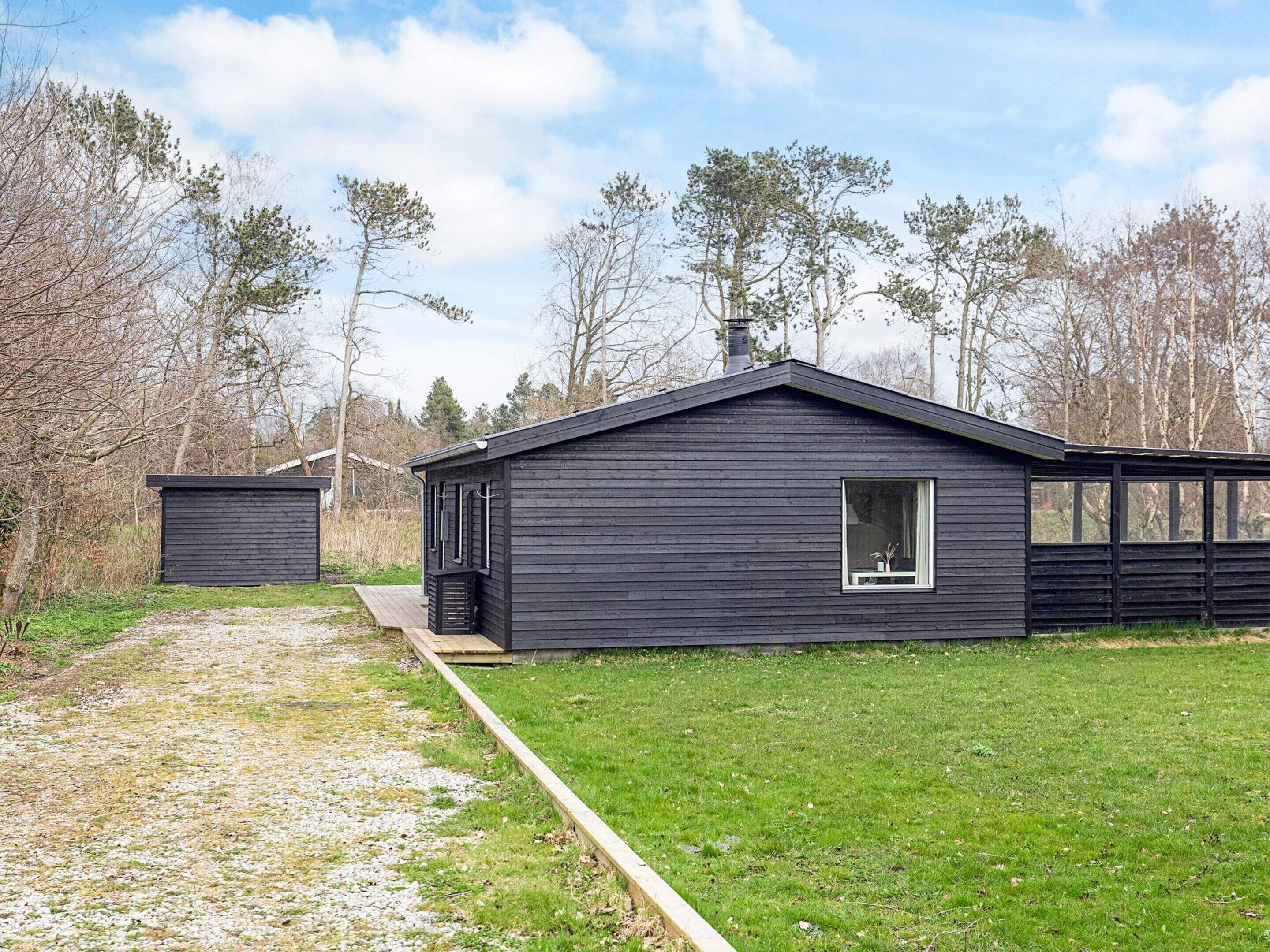A modern, dark wooden cabin with a large window, surrounded by a grassy area and trees.