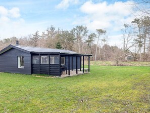 A small, dark wooden cabin with a porch, surrounded by a grassy field and trees. - 6 Person Holiday Home in Hals (Hals)