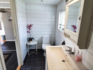 Une petite salle de bain moderne avec des murs recouverts de carreaux blancs, un lavabo blanc et des toilettes. On y trouve une petite table sur laquelle est posée une orchidée en pot et un récipient blanc.