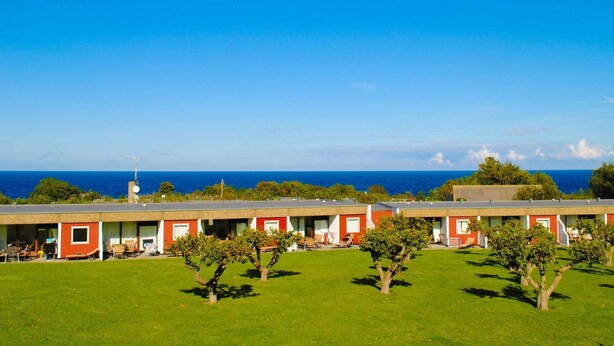 Eine Reihe von roten und weißen Motelgebäuden mit einer Rasenfläche und Bäumen im Vordergrund, mit Blick auf einen klaren, blauen Ozean.