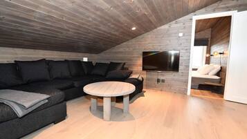 Un dormitorio tipo loft moderno con techo de madera, un televisor de pantalla plana grande montado en la pared, un sofá de color oscuro y una mesa redonda de madera.