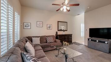 A living room with a sectional sofa, a glass coffee table, a dark wooden cabinet, a flat-screen TV, and a ceiling fan.