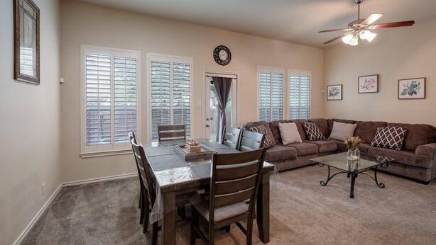 A spacious living room with a dining area, large windows, a ceiling fan, and a brown sofa with patterned cushions.