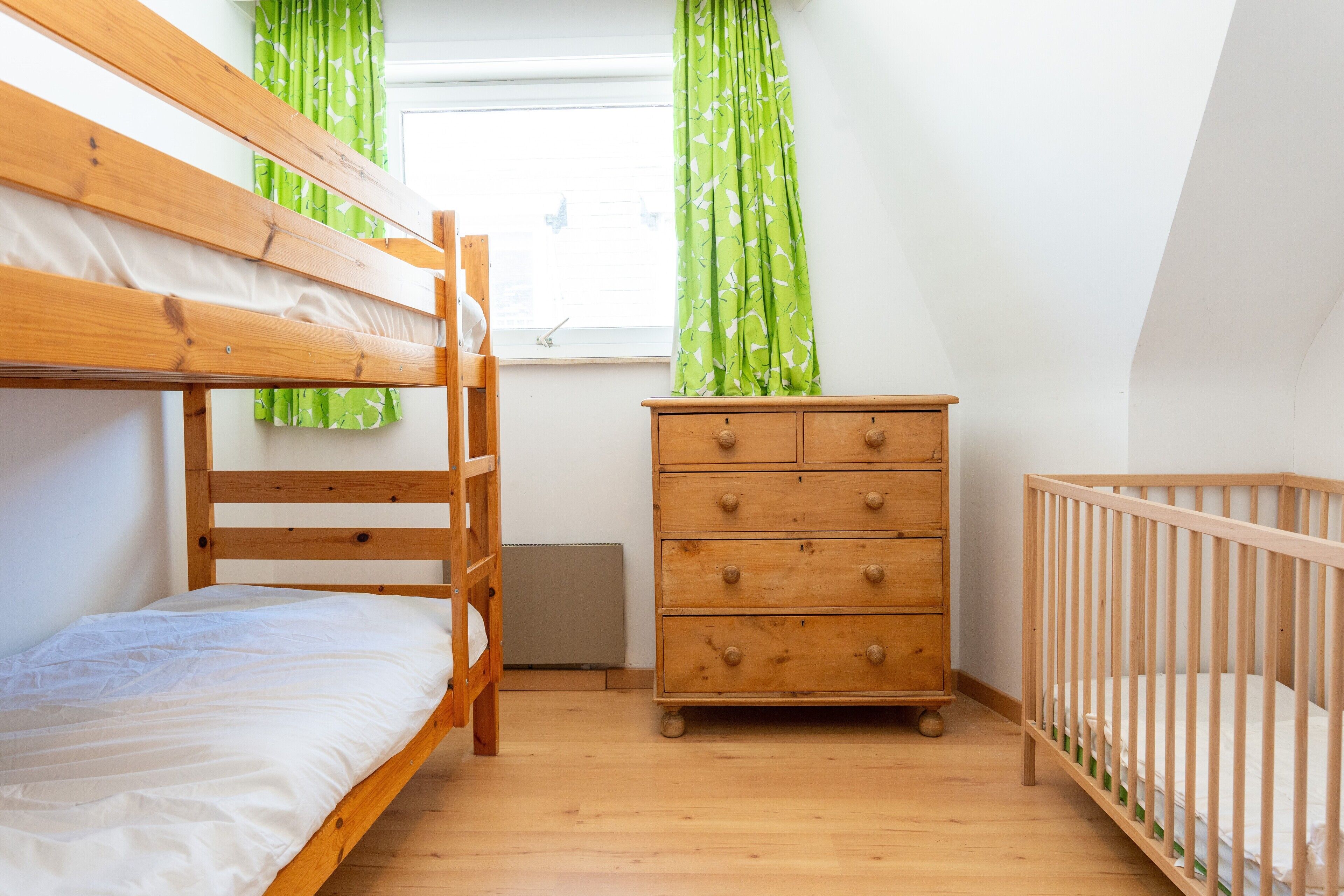A room with a bunk bed, a wooden dresser, a crib, and a window with green curtains.