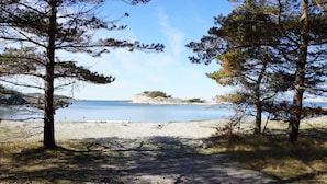 Un sendero bordeando la playa que lleva a una orilla arenosa, con árboles enmarcándola y mostrando una vista de un mar tranquilo y afloramientos rocosos a lo lejos.