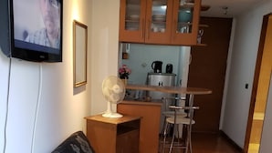 A compact living space with a sofa, a small table, a fan, a kitchenette, and a TV mounted on the wall.